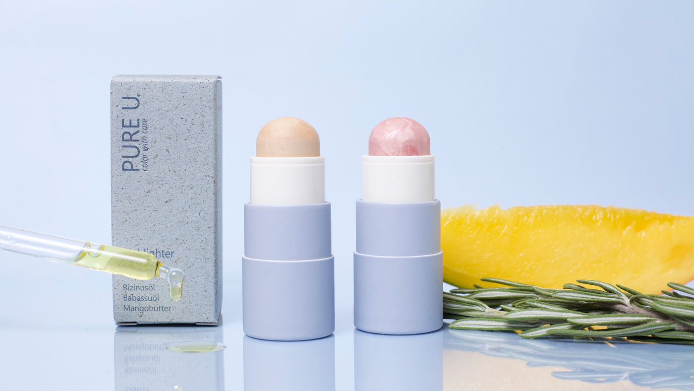 Die beiden Highlighter Balm Sticks von PURE U. vor einem blauen Hintergrund zusammen mit den Inhaltsstoffen Mango und Rosmarin